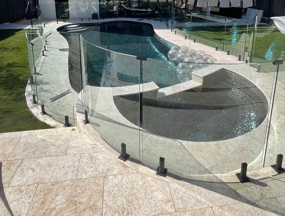 Finished pool and round spa behind frameless glass fencing, with travertine paving around them